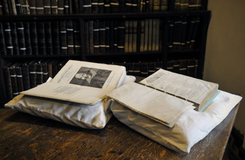 This picture shows the Durham First Folio next to a facsimile showing Shakespeare's portrait on the title page, which is now missing in the Durham copy. There are more old books on shelves in the background of the picture.