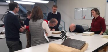 Picture showing a group of people in a conservation studio who are examining a damaged book. Another book lies closed on a cushion in the foreground.